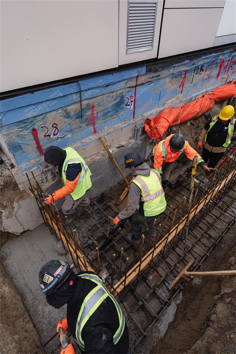 Blue waterproofing membrane on a foundation wall in an excavated trench, reinforcement staged for the footing